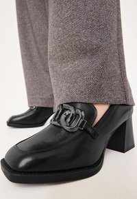 Černé kožené boty na podpatku se čtveratou špičkou a lesklým detailem s přezkou. V kombinaci s kalhotami s herringbone vzorem v purpurové a šedé barvě.