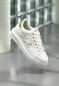 Adidași din piele albă cu accente aurii, având un logo texturat pe lateral, vârf rotund, talpă plată și un design clasic cu șireturi.