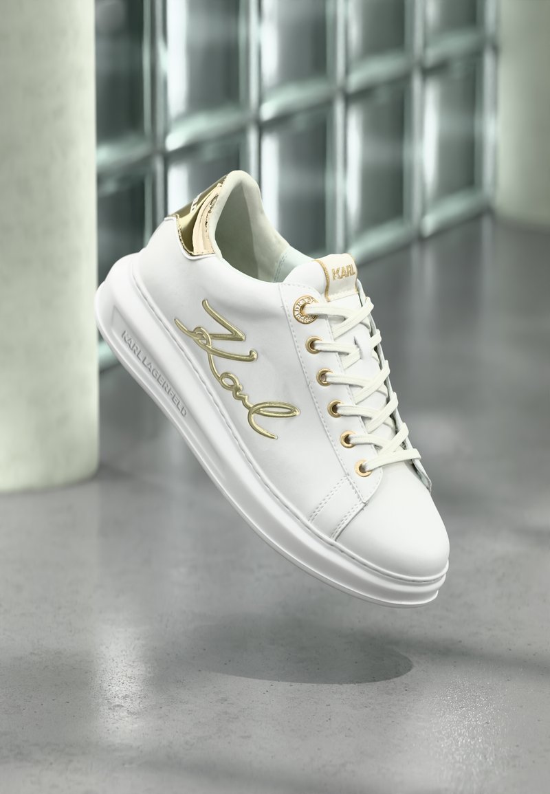 Adidași din piele albă cu accente aurii, având un logo texturat pe lateral, vârf rotund, talpă plată și un design clasic cu șireturi.
