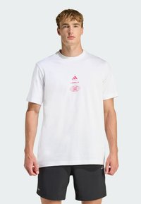 Camiseta de algodón blanca con cuello redondo, mangas cortas y detalles gráficos en rosa con el logo de Adidas. Combinada con pantalones cortos negros.