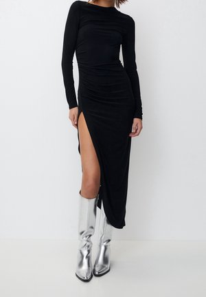 Femme portant une longue robe noire avec une haute fente et des bottes argentées montantes, se tenant devant un fond blanc uni.