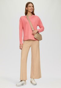 Roze gebreide trui met een ronde halslijn, brede beige broek en lichtgekleurde laarzen. Het model houdt een bruine schoudertas vast.