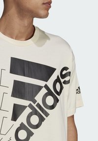 Camiseta de manga corta color crema con gran logo negro de adidas y acentos gráficos en la parte frontal, además de un pequeño logo en la manga.
