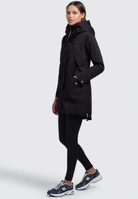 Parka noire avec capuche, fermeture éclair à l'avant, deux grandes poches et un ourlet à cordon, portée sur des leggings noirs et des chaussures de sport bleues.