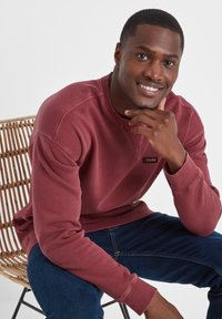 Man seated on wicker chair wearing a maroon sweatshirt and blue jeans, smiling with hand resting on chin against a white background.