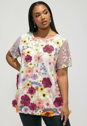 Donna che indossa una camicetta bianca trasparente con fiori ricamati colorati in rosa, viola, giallo e verde, abbinata a pantaloni neri.