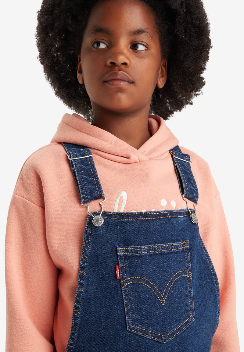 Denim-Latzhose mit verstellbaren Trägern, einer Brusttasche mit einem roten Levi's-Label, getragen über einem rosa Kapuzenpullover. Dunkelblauer Stoff, glatte Textur.