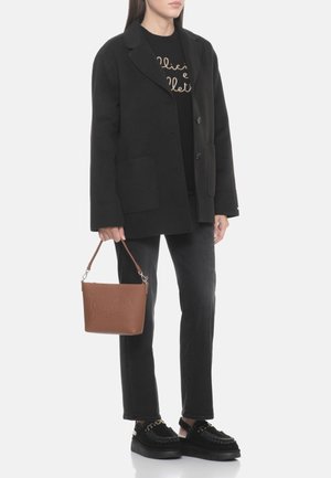 Donna che indossa un blazer nero, pantaloni neri, scarpe nere e tiene in mano una borsa di pelle marrone con testo in rilievo.