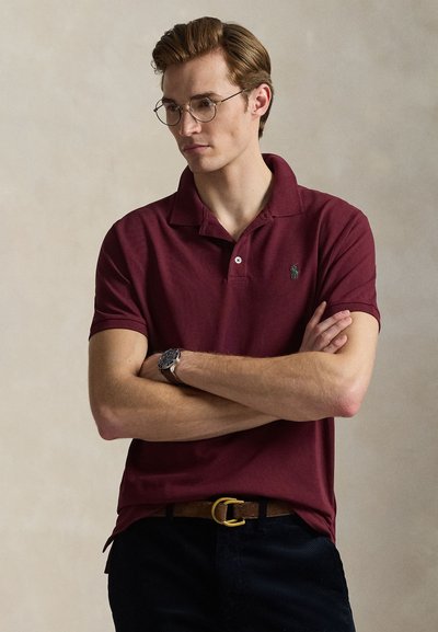 Vínová polokošile s krátkými rukávy, kontrastním zeleným logem, v kombinaci s tmavými kalhotami a hnědým pásem se zlatou sponou.