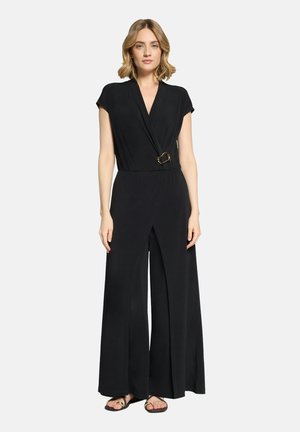 Woman wearing a black short-sleeve wrap jumpsuit with wide legs and a gold oval buckle detail at the waist, paired with black sandals.