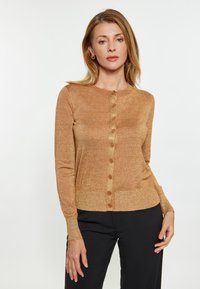 Goldfarbener, glitzernder Cardigan mit Rundhalsausschnitt, sieben Vorderknöpfen und gerippten Bündchen. Enganliegend, weicher Stoff, kombiniert mit schwarzen Hosen.