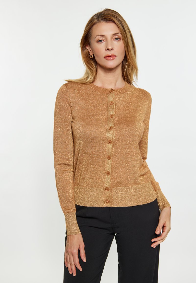 Goldfarbener, glitzernder Cardigan mit Rundhalsausschnitt, sieben Vorderknöpfen und gerippten Bündchen. Enganliegend, weicher Stoff, kombiniert mit schwarzen Hosen.