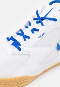White athletic shoe featuring blue laces and logo accents, textured mesh upper, smooth synthetic overlays, and a gum rubber outsole with a geometric pattern.