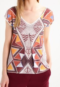 Top a maniche corte con un motivo geometrico multicolore nei toni del marrone, giallo e blu. Design a V con una vestibilità rilassata e tessuto a trama.