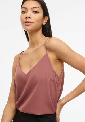 Žena s dlhými tmavými vlasmi má na sebe fialovohnedý bezrukávový top s výstrihom do V a čierne nohavice, pózuje s jednou rukou na boku na bielom pozadí.