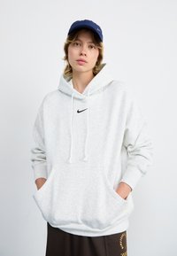 Grijze hoodie met een voorzak, een capuchon met trekkoord en een klein zwart logo. Gecombineerd met een marineblauwe pet en een donkerbruine rok.