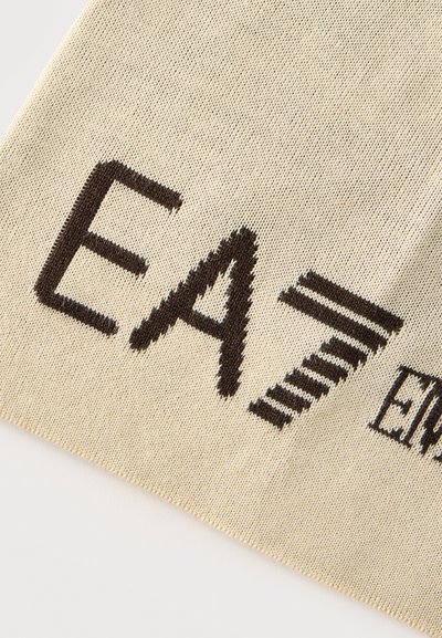 Gros plan sur un tissu beige avec un texte tricoté noir montrant "EA7" et un texte partiel plus petit sur le côté droit.