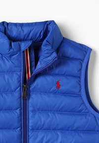 Blå vadderad väst med hög krage, dragkedja fram och röd broderad logotyp. Har ett horisontellt quiltat mönster och kontrasterande inredning.