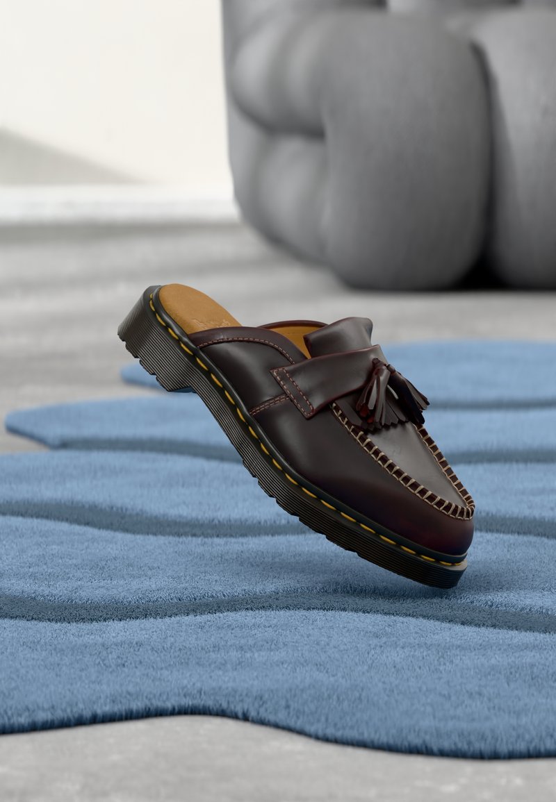 Dunkelbrauner Leder-Slipper mit Quasten und gelb gestepptem Absatz auf einem strukturierten blauen Teppich, im Hintergrund unscharf ein graues Kissen.