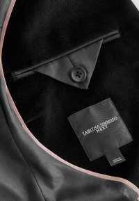 Intérieur d'une veste en tissu noir avec une doublure en satin rose, un bouton unique, et une étiquette portant la mention "TABITHA SIMMONS NEXT" en texte blanc.