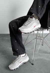 Stříbrné tenisky Nike Air Max 97 mají vlnitý design, červené logo a viditelnou vzduchovou jednotku, doplněné černými džínami s bílými puntíky.