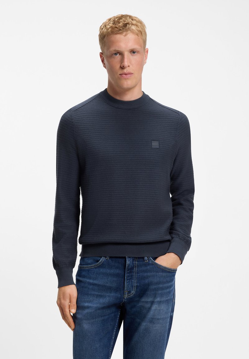Jeune homme aux cheveux blonds courts, portant un pull texturé bleu marine foncé et un jean bleu, debout avec une main dans la poche sur fond blanc.