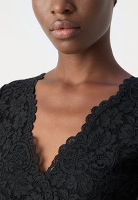 Haut en dentelle noire avec un décolleté en V profond, présentant des motifs floraux complexes et des bords festonnés, mettant en avant un tissu texturé et un design délicat.