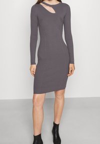 Robe en maille côtelée grise avec manches longues, présentant une découpe au niveau de l'encolure. Coupe slim, longueur genoux, portée avec des bottines noires.