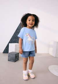 Chemise à manches courtes bleu clair avec un motif de licorne, associée à un short en denim et des baskets pastel avec des détails en dentelle. Fond neutre.