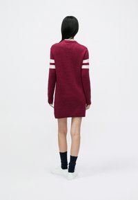 Bordeauxrotes Strickkleid mit langen Ärmeln, weißen Streifen auf den Schultern, gerippten Bündchen und Saum. Kombiniert mit marineblauen Socken und weißen Turnschuhen.