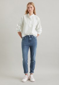 Witte blouse met lange mouwen en een knoopsluiting, gecombineerd met lichtblauwe tapered jeans en witte sneakers met roze accenten.