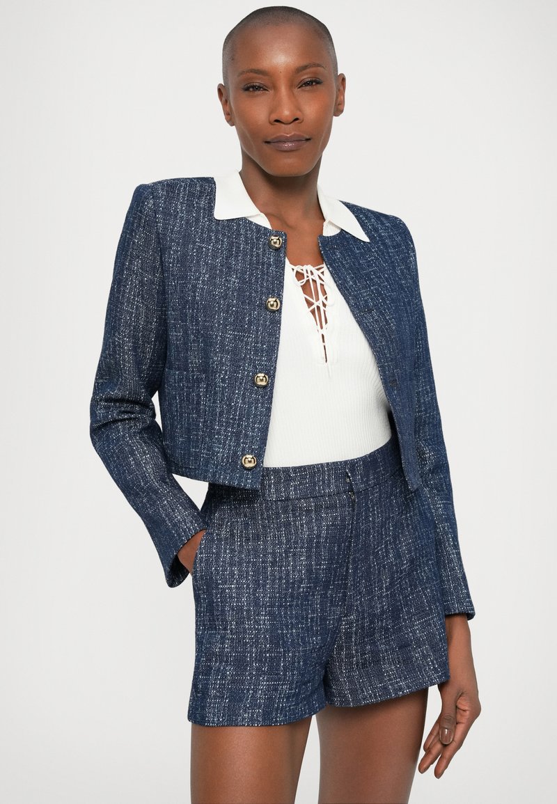 Marineblauer Tweed-Cropped-Jacke mit goldenen Knöpfen, kombiniert mit passenden Shorts. Darunter befindet sich ein weißes Oberteil mit Kragen und einer Schnürung am Hals.