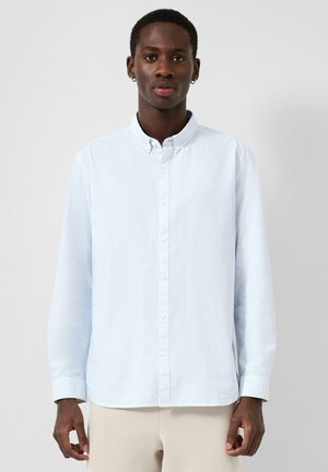 Junger Mann trägt hellblaues Hemd mit Knöpfen und beige Hose, steht vor einem einfarbigen hellgrauen Hintergrund und blickt nach vorne.