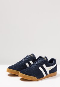 Baskets en daim bleu marine avec deux rayures blanches, une semelle en caoutchouc gomme et des accents dorés sur la languette et sur le côté. Design athlétique classique.