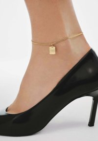 Tobillo con una delgada cadena de oro que presenta un pequeño charm rectangular, combinado con un zapato de tacón alto de charol negro.