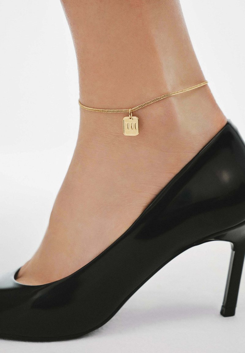 Tobillo con una delgada cadena de oro que presenta un pequeño charm rectangular, combinado con un zapato de tacón alto de charol negro.