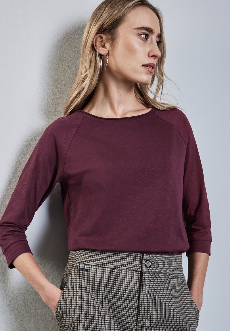 Burgundy pitkähihainen paita pyöreällä pääntiellä, yhdistettynä houndstooth-kuosiset housut. Sisältää rentoa istuvuutta ja pehmeää tekstuuria.