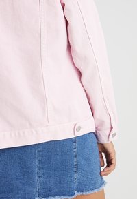 Chaqueta de mezclilla rosa con cierres a presión plateados, costuras visibles y un diseño recortado, combinada con una falda de mezclilla azul con bordes deshilachados.