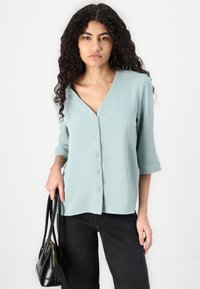 Blusa verde chiaro con bottoni in un taglio rilassato, caratterizzata da un scollo a V e maniche a tre quarti, abbinata a pantaloni neri e una piccola borsa.