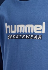 T-shirt en coton bleu avec des accents bleu marine. Comprend un grand logo blanc "hummel" et le mot "SPORTSWEAR" en bleu marine en dessous.
