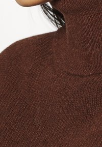 Brązowy sweter z golfem wykonany z dzianiny, o strukturalnym, ryflowanym wzorze i dopasowanym kroju, podkreślający miękki, ciepły materiał i bezszwową konstrukcję.