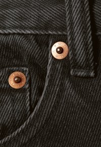 Tecido de ganga preta com padrão espinha de peixe, com rebites e costuras de cor cobre ao longo das bordas. Close-up do detalhe do bolso.