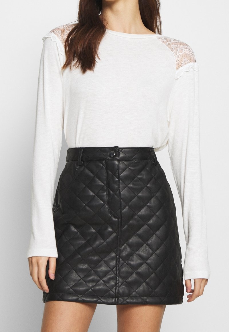 Donna che indossa una maglietta bianca a maniche lunghe con spalle in pizzo e una minigonna nera in pelle trapuntata, in piedi di fronte a uno sfondo neutro.