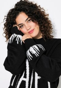Schwarzer Pullover mit weißem, tropfendem Muster, gerippten Bündchen und weichem, strukturiertem Stoff. Modell posiert mit Händen nahe dem Gesicht und präsentiert das Design.