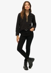 Haut noir en polaire avec fermeture éclair contrastante dorée, associé à un jean noir ajusté et des bottes à lacets noires. Texture lisse, design décontracté.