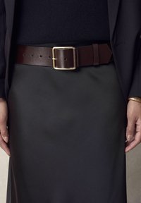 Cintura in pelle marrone con una fibbia rettangolare dorata, indossata sopra una gonna in tessuto scuro, con una superficie liscia e cinque fori regolabili.