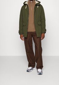 Grön vadderad jacka med huva, brun tröja och bruna byxor. Funktioner inkluderar dragkedja framtill och framfickor. Sneakers är grå och vita.