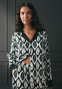 Femeie cu păr lung și închis la culoare, îmbrăcată în pijamale cu un model alb-negru ce prezintă mâini cu motive de inimi, stând în fața unui perete închis la culoare.