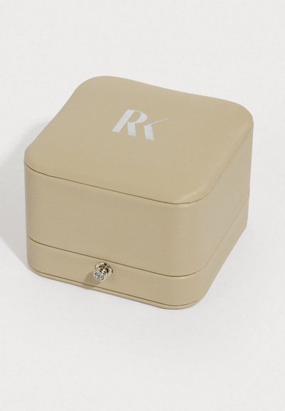 Štvorcová šperkovnica z jemného béžového materiálu, so strieborným logom a okrúhlym kovovým uzáverom vpredu. Hladká textúra, minimalistický dizajn.