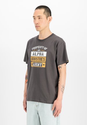 Man met kort haar die een donkergrijs "Alpha Industries Army" T-shirt en lichtblauwe jeans draagt, staand tegen een witte achtergrond.